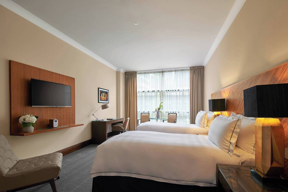 Une chambre d’hôtel avec deux lits, un téléviseur fixé au mur, un bureau avec une chaise, une petite table avec un vase et une fenêtre avec des rideaux.