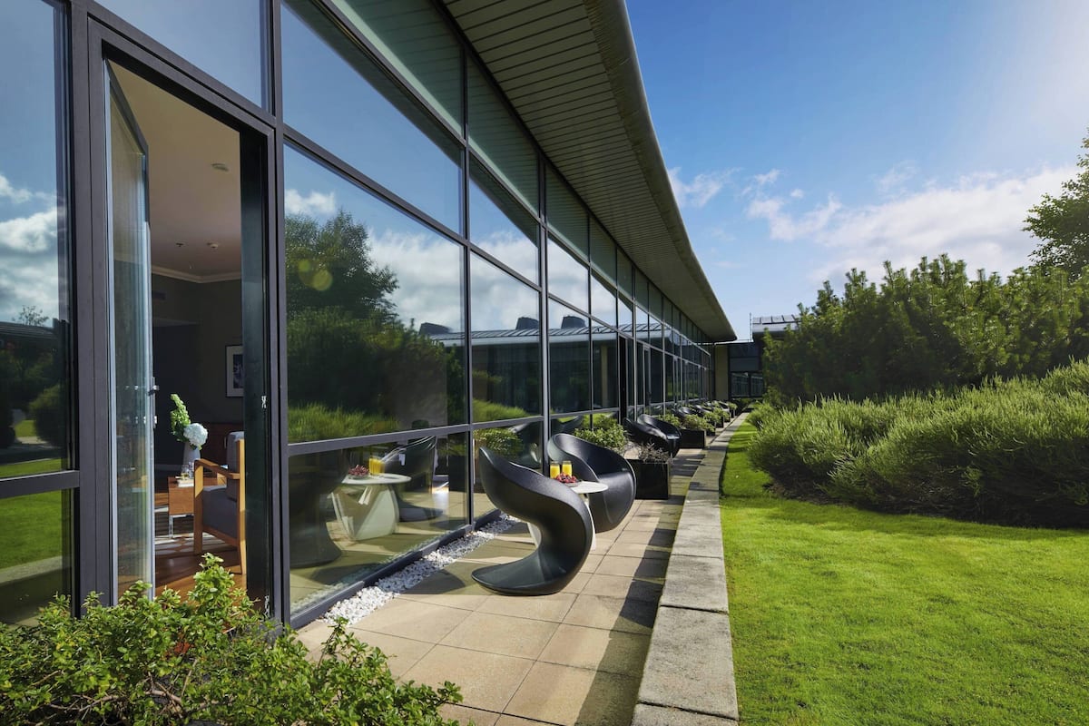 Un bâtiment moderne doté de grandes fenêtres en verre, d’un espace extérieur aménagé avec des chaises et des tables, et d’une pelouse bien entretenue.