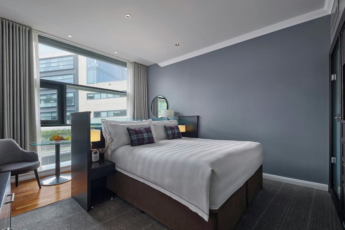 Une chambre d’hôtel moderne avec un grand lit, une table de chevet avec une lampe, un fauteuil et une vue sur la ville grâce à de grandes fenêtres.