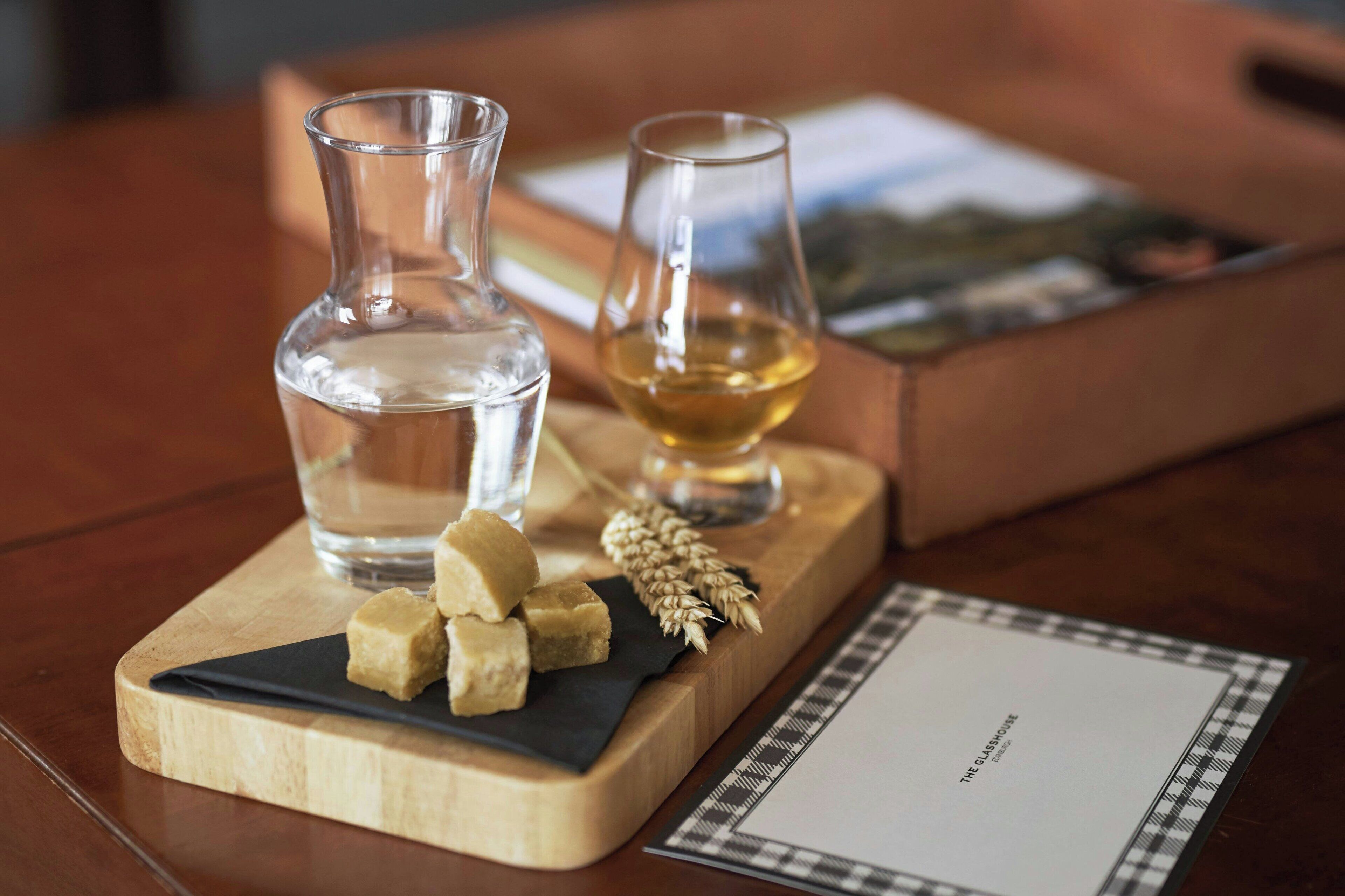 A wooden board with a bottle of water, a glass of whiskey, and some cheese cubes on a table.