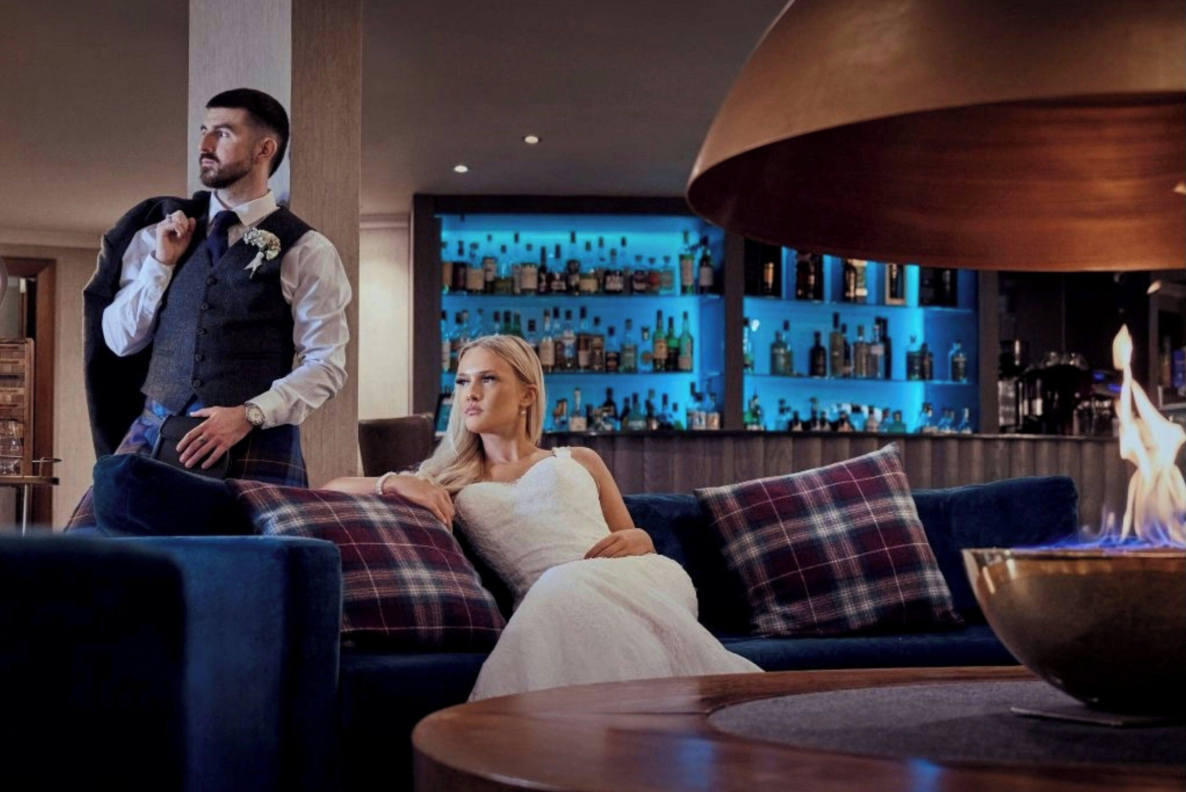 A man in a vest and tie standing beside a woman in a white dress sitting on a blue sofa with plaid cushions. A bar with shelves of bottles is in the background.