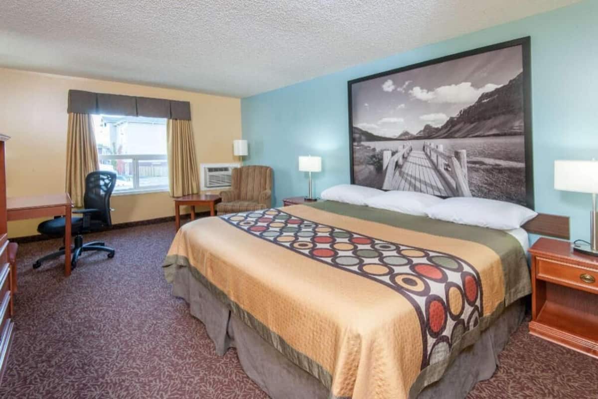 Habitación de hotel con cama, escritorio, silla, ventana con cortinas y un cuadro grande en la pared.