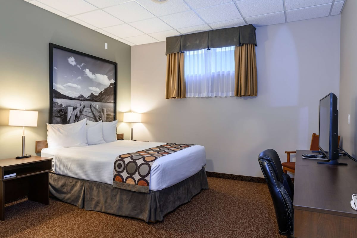 A hotel room with a bed, two bedside lamps, a desk with a computer, a chair, and a large framed picture on the wall.