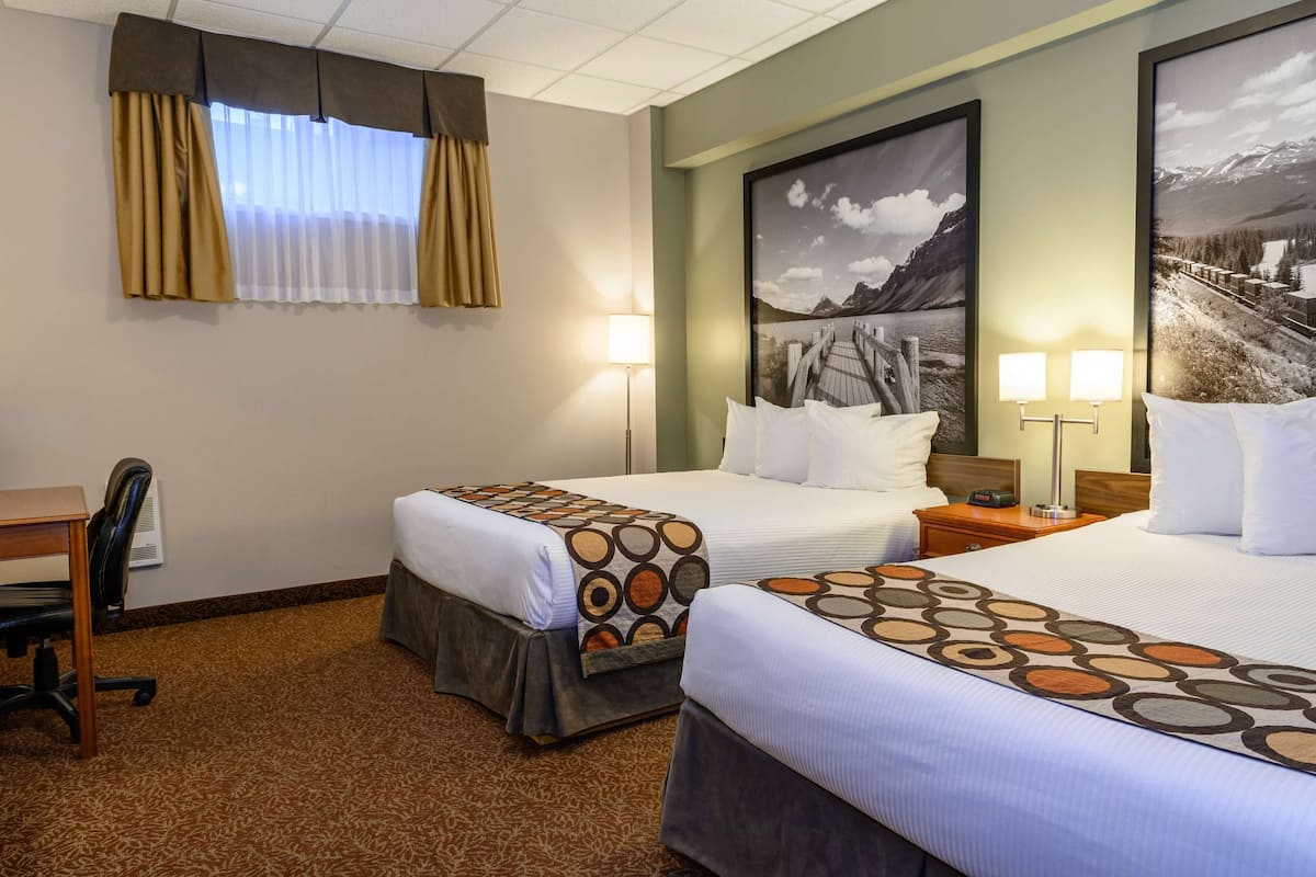 A hotel room with two beds, a desk, and a chair. There are two wall-mounted lamps and a large framed picture on the wall.