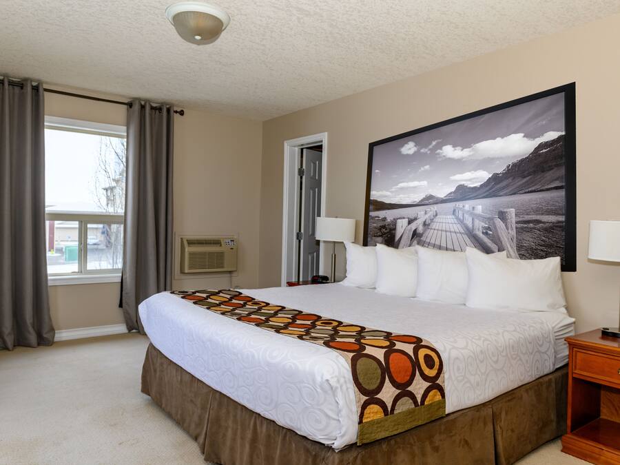 Ein Hotelzimmer mit einem großen Bett, einem Fenster mit Vorhängen, einer Wand-Klimaanlage, einem gerahmten Bild und einem Nachttisch mit Lampe.
