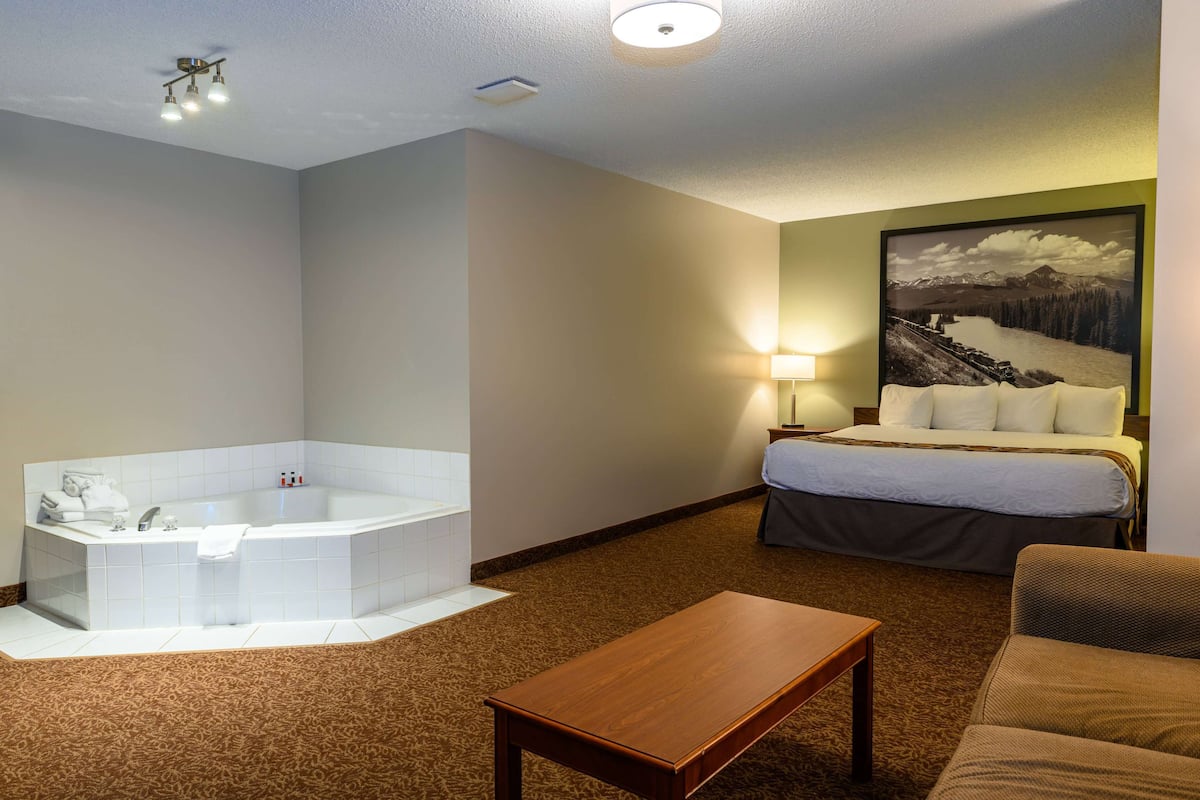 A hotel room with a large bathtub, a bed with a green wall, a lamp, and a painting of a mountain landscape.