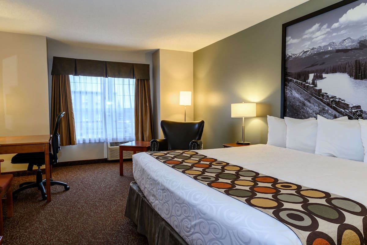 A hotel room with a large bed, a desk, a chair, a window with curtains, and a wall-mounted lamp.