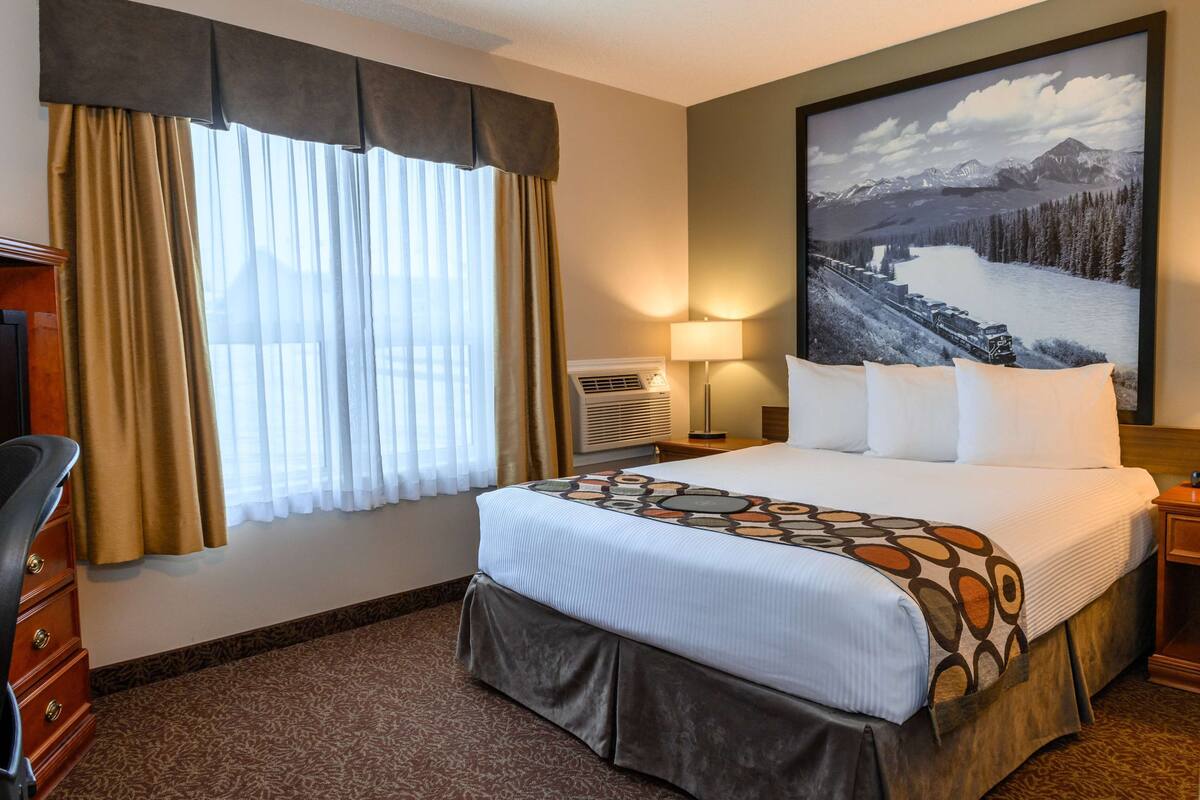 A hotel room with a large bed, a desk, a chair, a lamp, and a painting of a mountainous landscape.