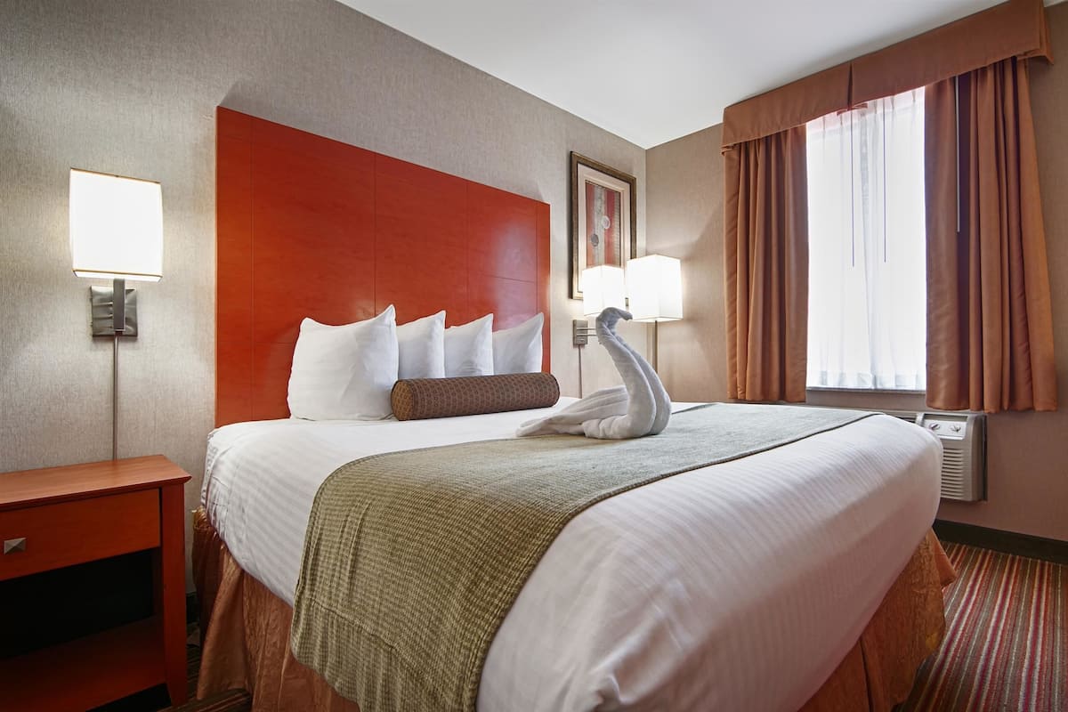 A hotel room with a large bed, a desk, a chair, a wall-mounted lamp, and a window with curtains.