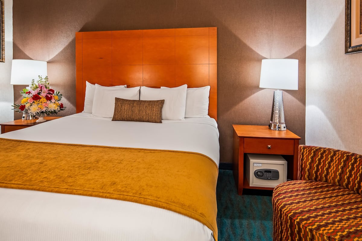 A hotel room with a large bed, a bedside table with a lamp, a small table with a vase of flowers, and a patterned armchair.
