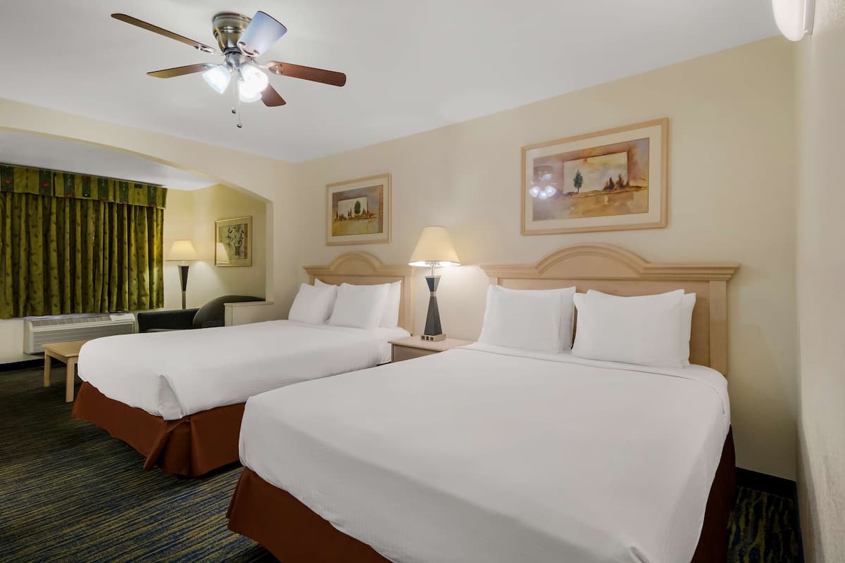 A hotel room with two beds, a ceiling fan, a desk, a chair, and artwork on the walls.
