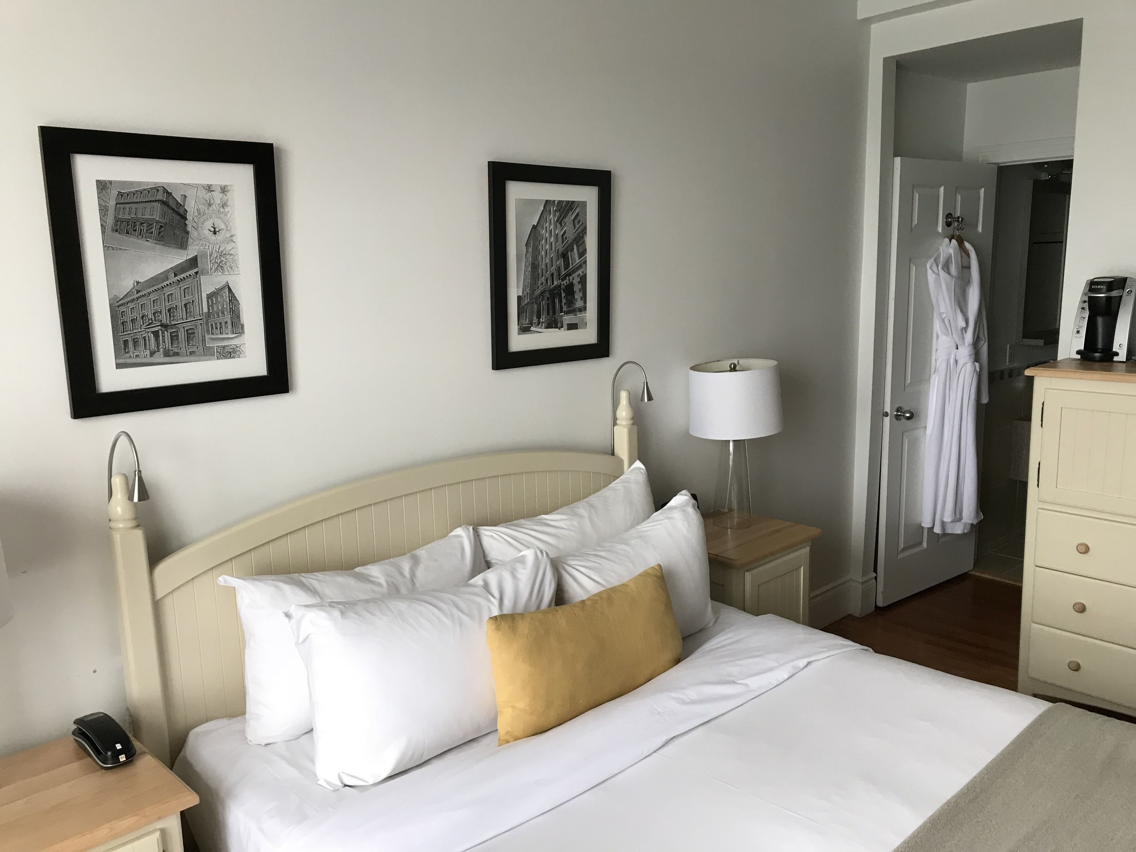 Una habitación de hotel con una cama, dos cuadros en la pared, una lámpara de noche, una bata colgada en la puerta y una cafetera sobre la mesita de noche.