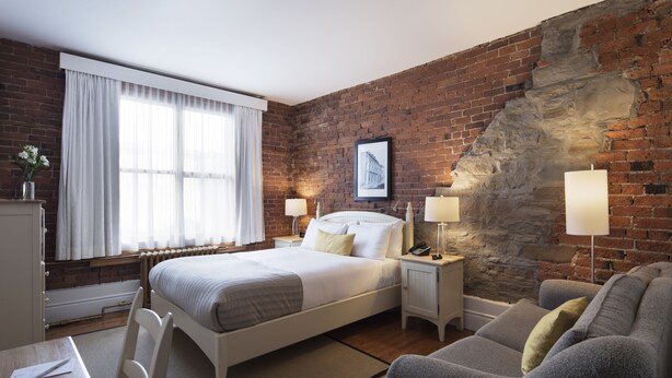 A hotel room with a brick wall, a large bed, two bedside tables with lamps, a gray sofa, and a window with sheer curtains. - Auberge Saint-Pierre (Québec City)