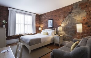 Een hotelkamer met een bakstenen muur, een groot bed, twee nachtkastjes met lampen, een grijze bank en een raam met dunne gordijnen.