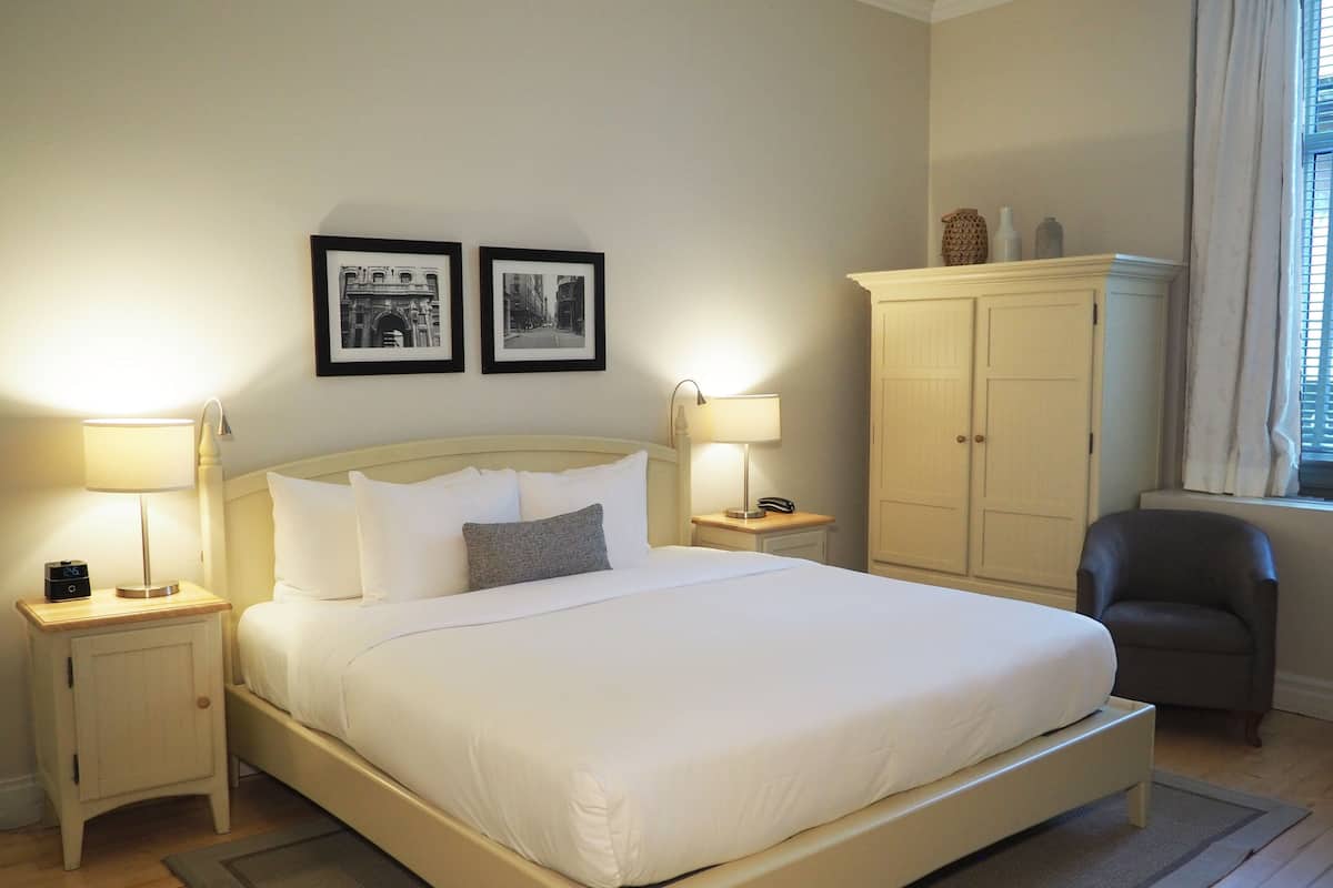 A neatly made bed with white linens, two bedside lamps, a nightstand with a clock, a wardrobe, and two framed pictures on the wall.