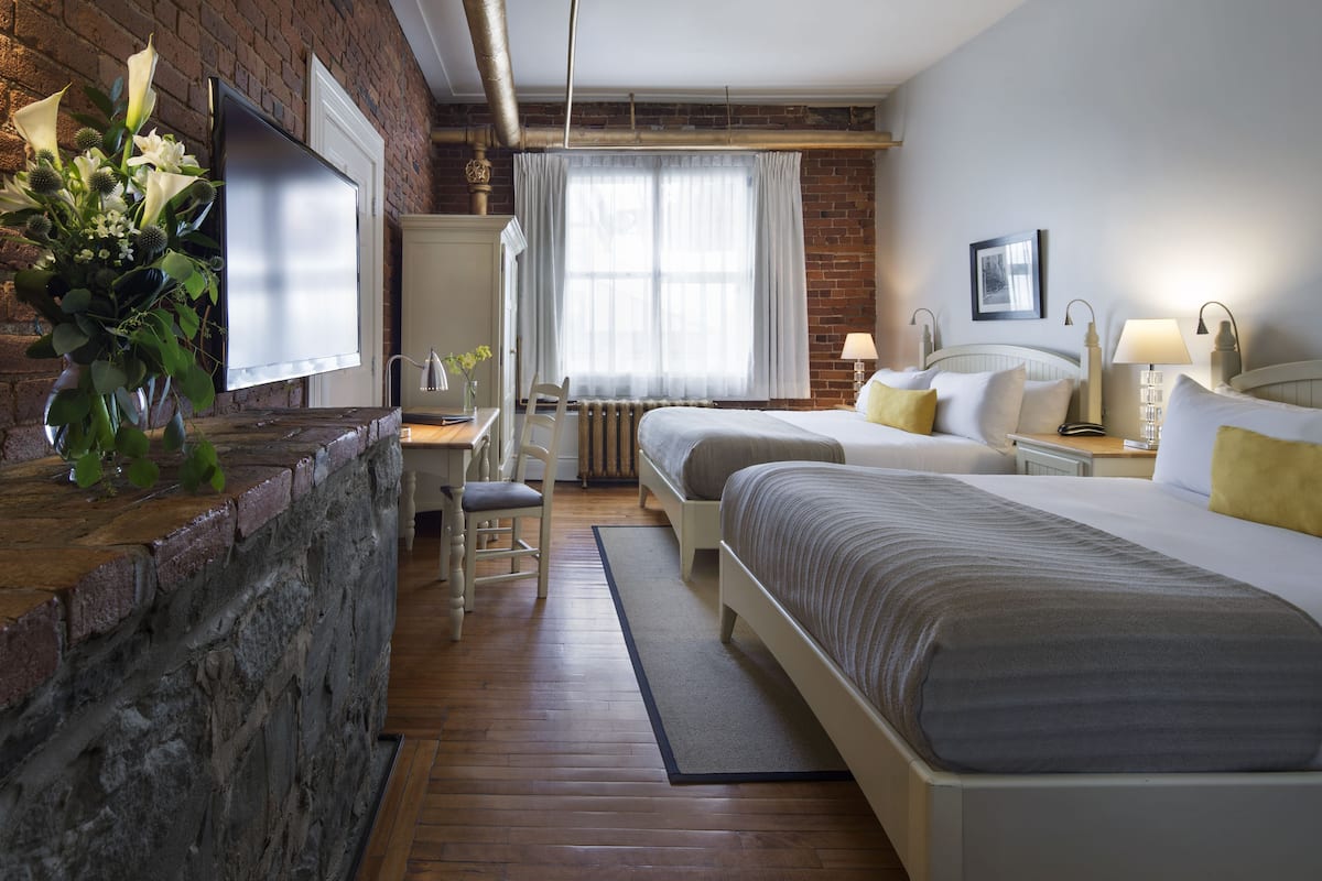 A hotel room with two beds, a desk, a chair, a radiator, and a TV mounted on a brick wall.