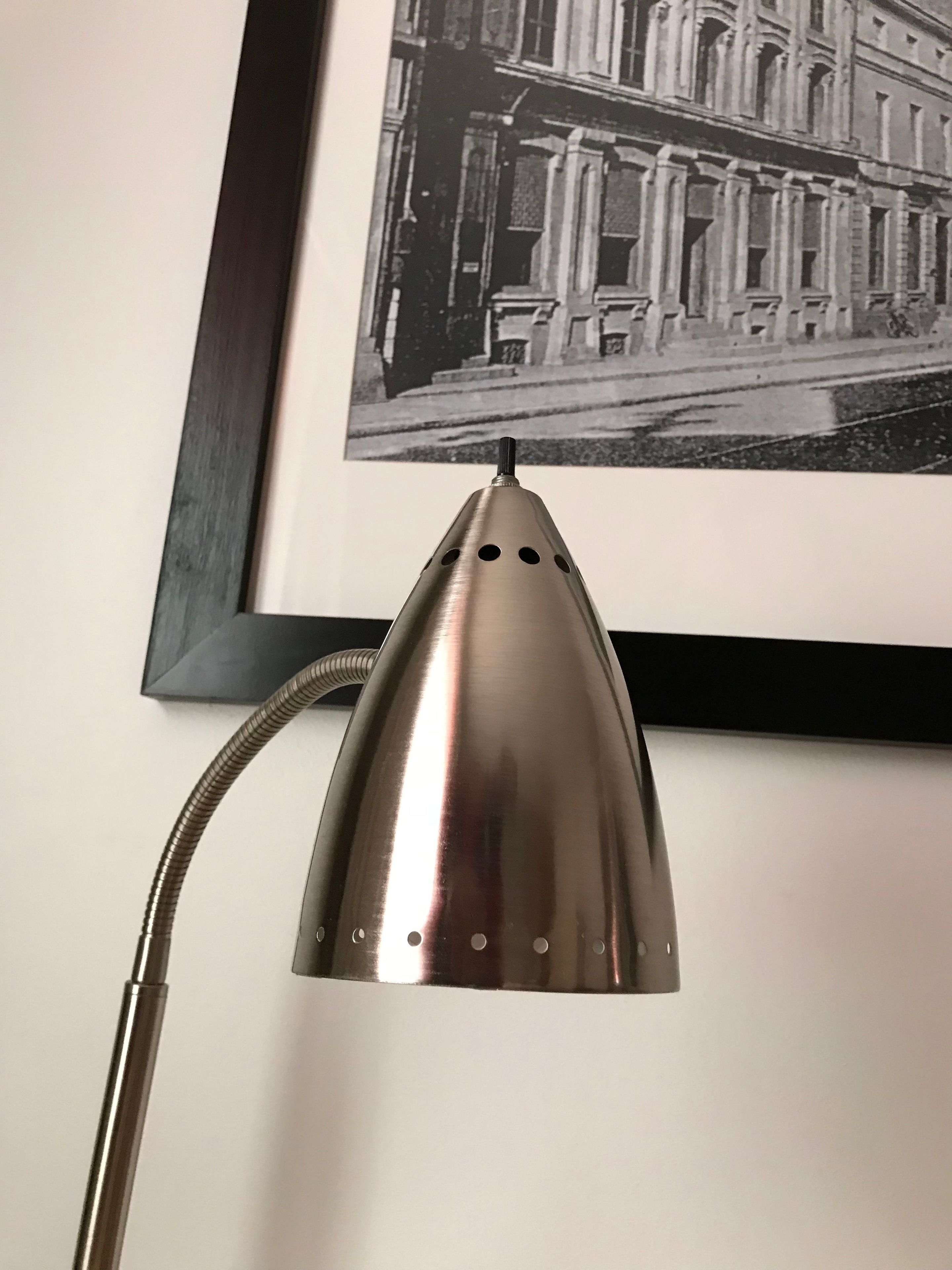 A metallic desk lamp with a flexible neck, positioned in front of a framed black and white photograph of a classical building.