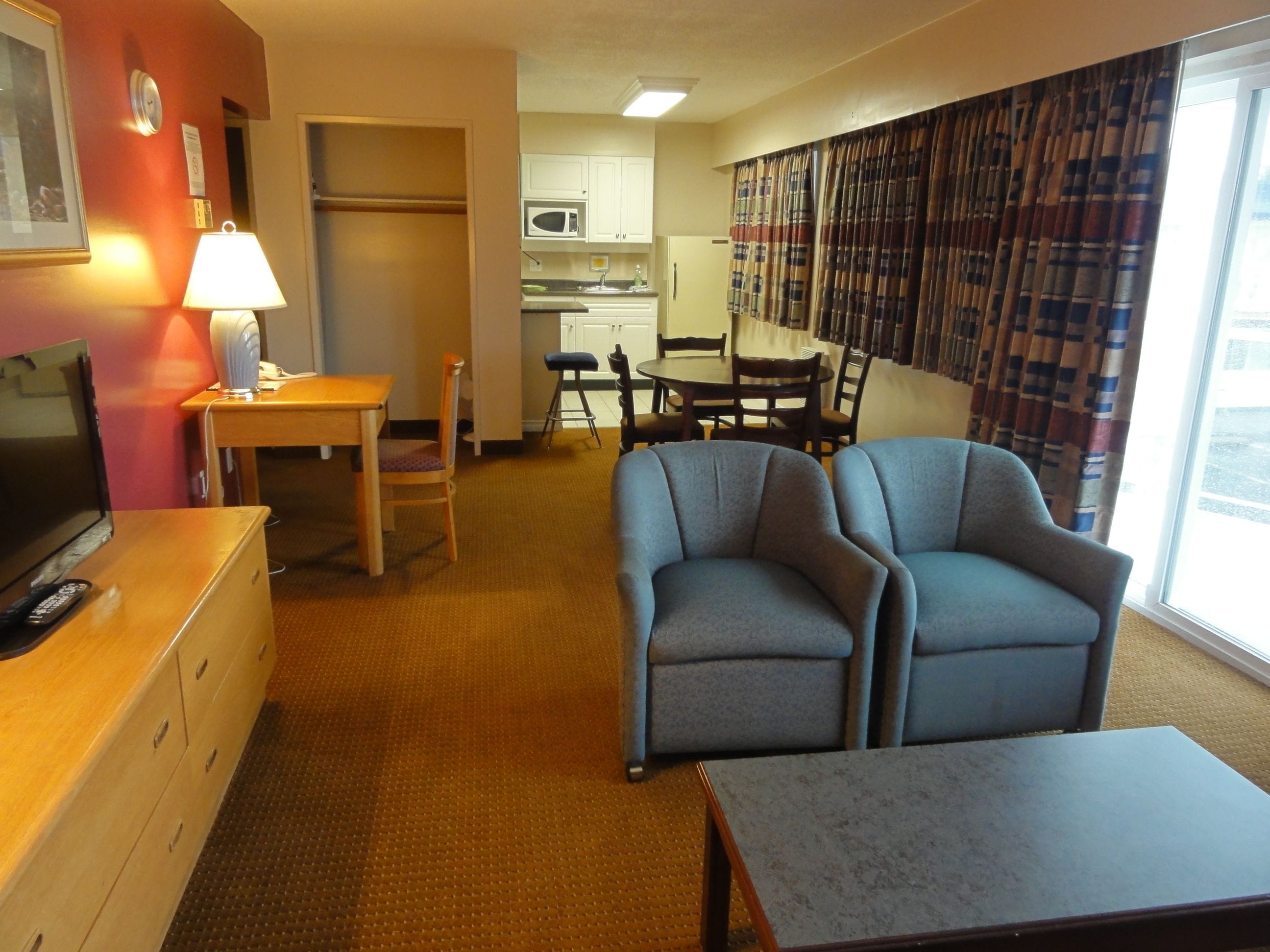 A hotel room with a TV, a desk with a lamp, a dining area with chairs, a sofa, and a kitchen area with a microwave.