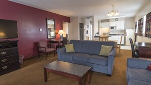 A living room with a sofa, coffee table, and TV. There is a kitchen in the background with cabinets and appliances. - Port Augusta Inn and Suites (Comox)