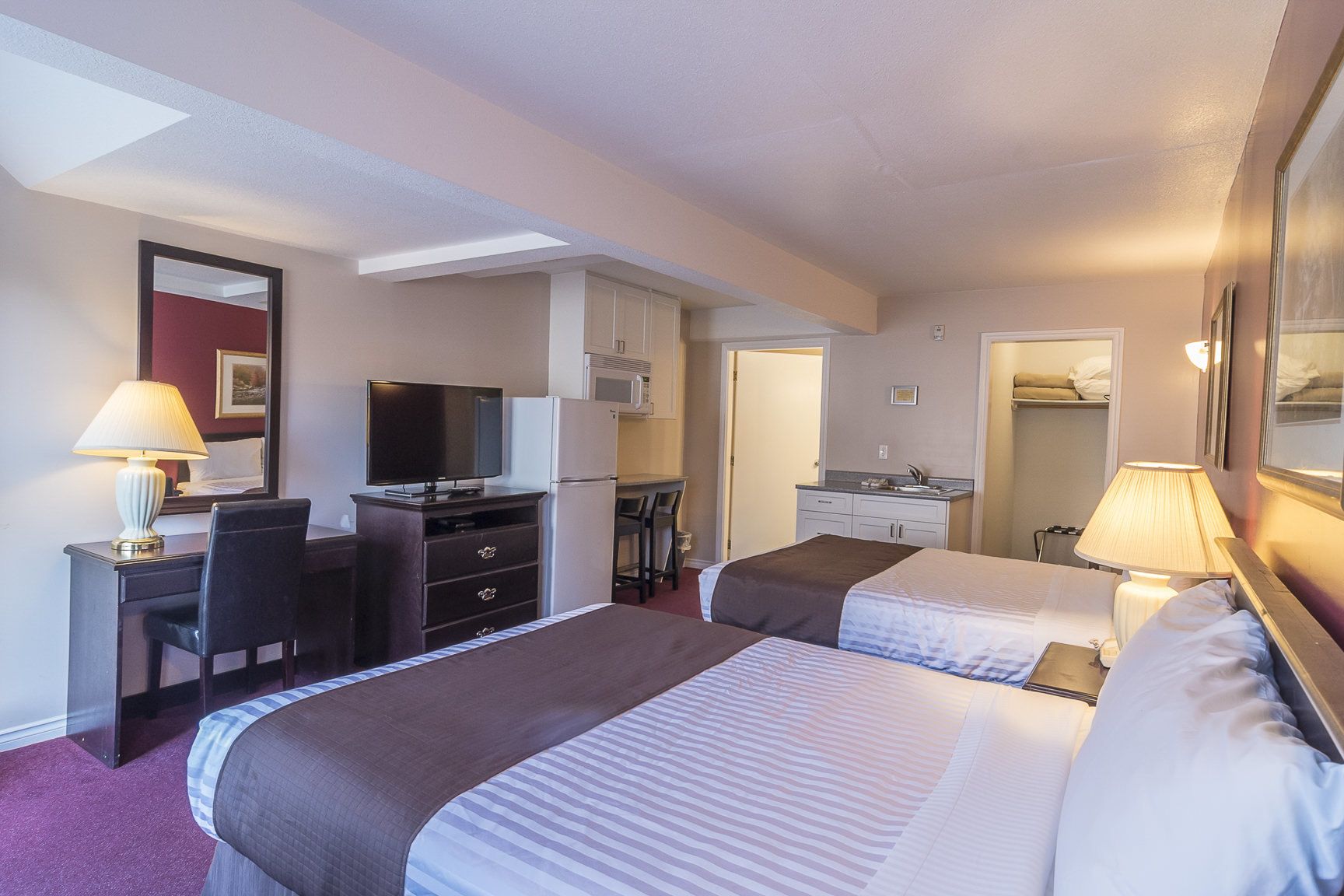 A hotel room with two beds, a desk with a television, a refrigerator, a microwave, a small kitchenette, and a bathroom.