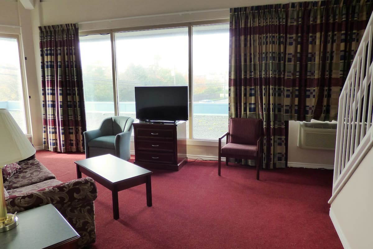 Habitación de hotel con televisión, sofá, sillón, mesa y ventanas grandes con cortinas.
