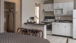 A compact kitchen with white cabinets, a microwave, stove, and a refrigerator. There is a dining area with a table and chairs. - Port Augusta Inn and Suites (Comox)