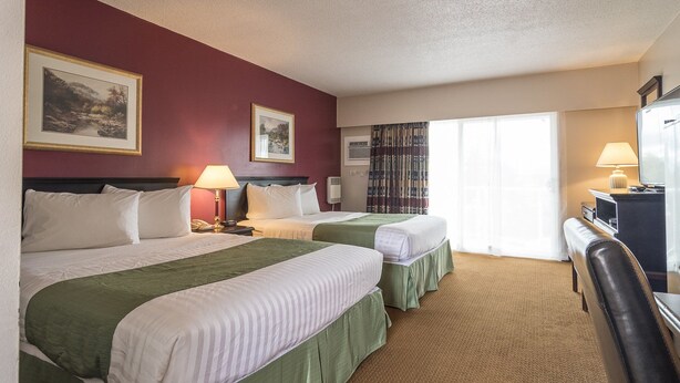 A hotel room with two beds, a desk, a chair, and a large window with curtains. - Port Augusta Inn and Suites (Comox)