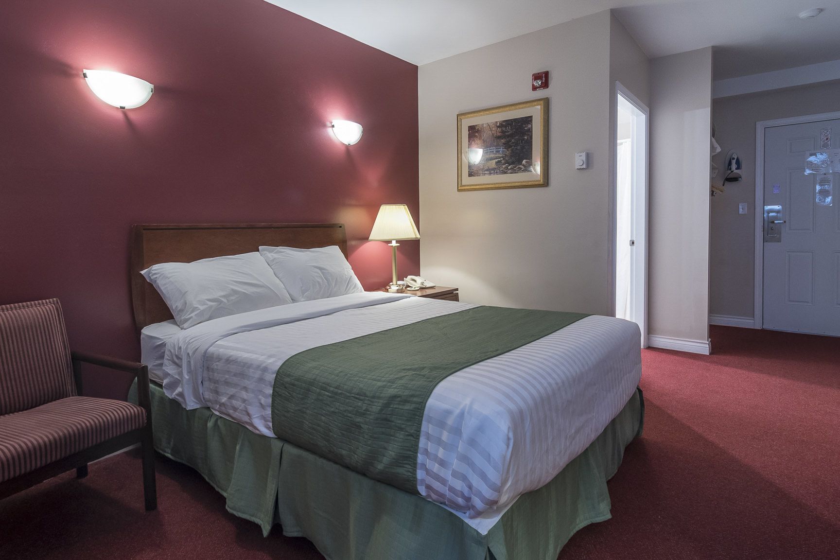 Habitación de hotel con una cama, una silla, una mesita de noche, una lámpara, un cuadro enmarcado en la pared y una puerta.