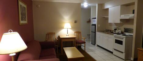 A compact living space with a sofa, a small table, a lamp, a chair, a kitchenette with a stove, microwave, and refrigerator.