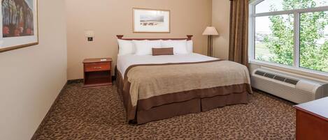 A hotel room with a large bed, a nightstand, a lamp, a framed picture on the wall, and a window with a view of greenery.