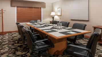 A conference room with a long table, black leather chairs, a patterned carpet, a window with blinds, a lamp, and a framed picture on the wall.