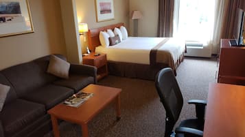 A hotel room with a bed, a sofa, a desk, and a chair. There is a lamp, a picture frame, and a window with curtains.