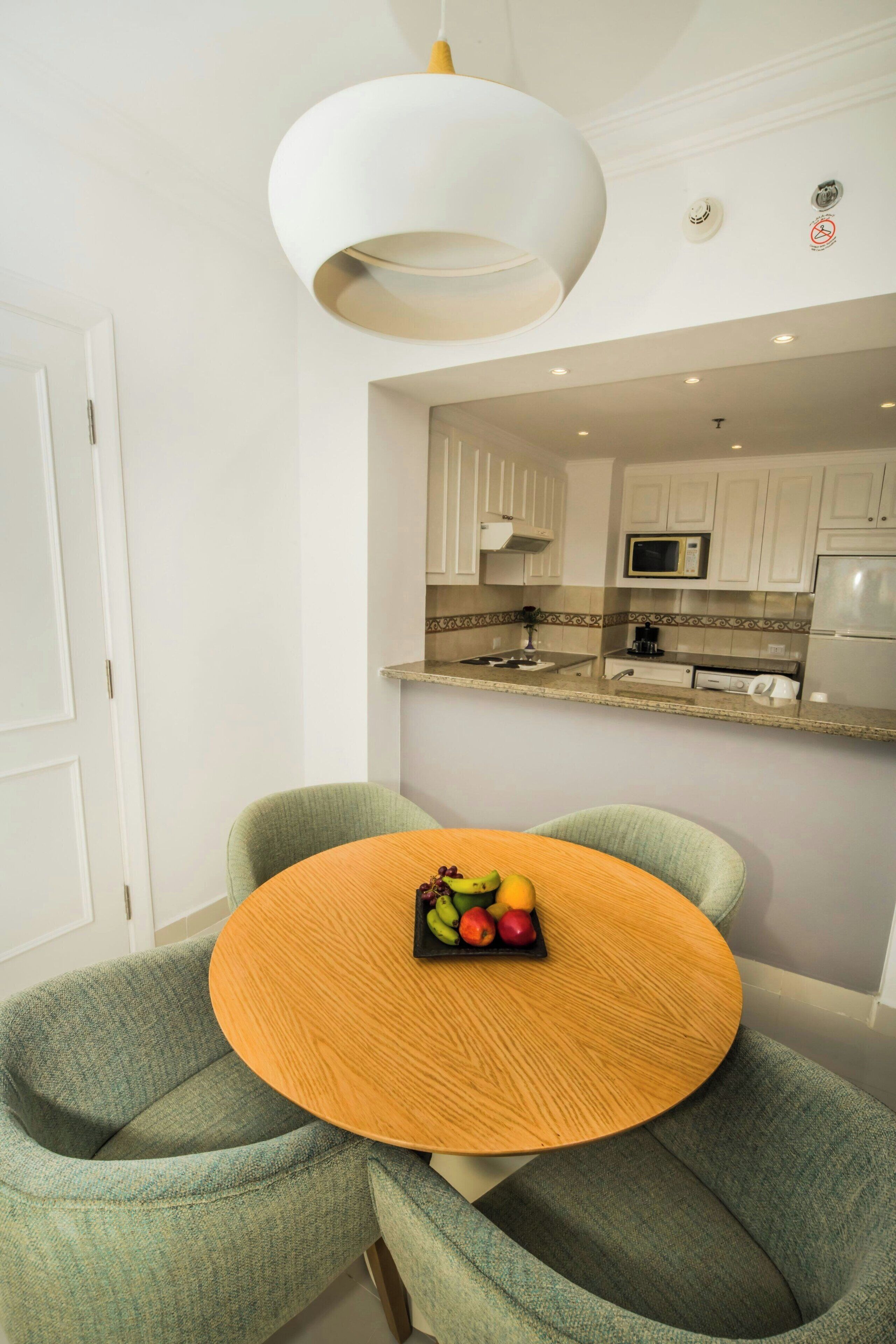 A modern kitchen with white cabinets, a microwave, and a refrigerator. There is a dining area with a round wooden table and two green chairs.