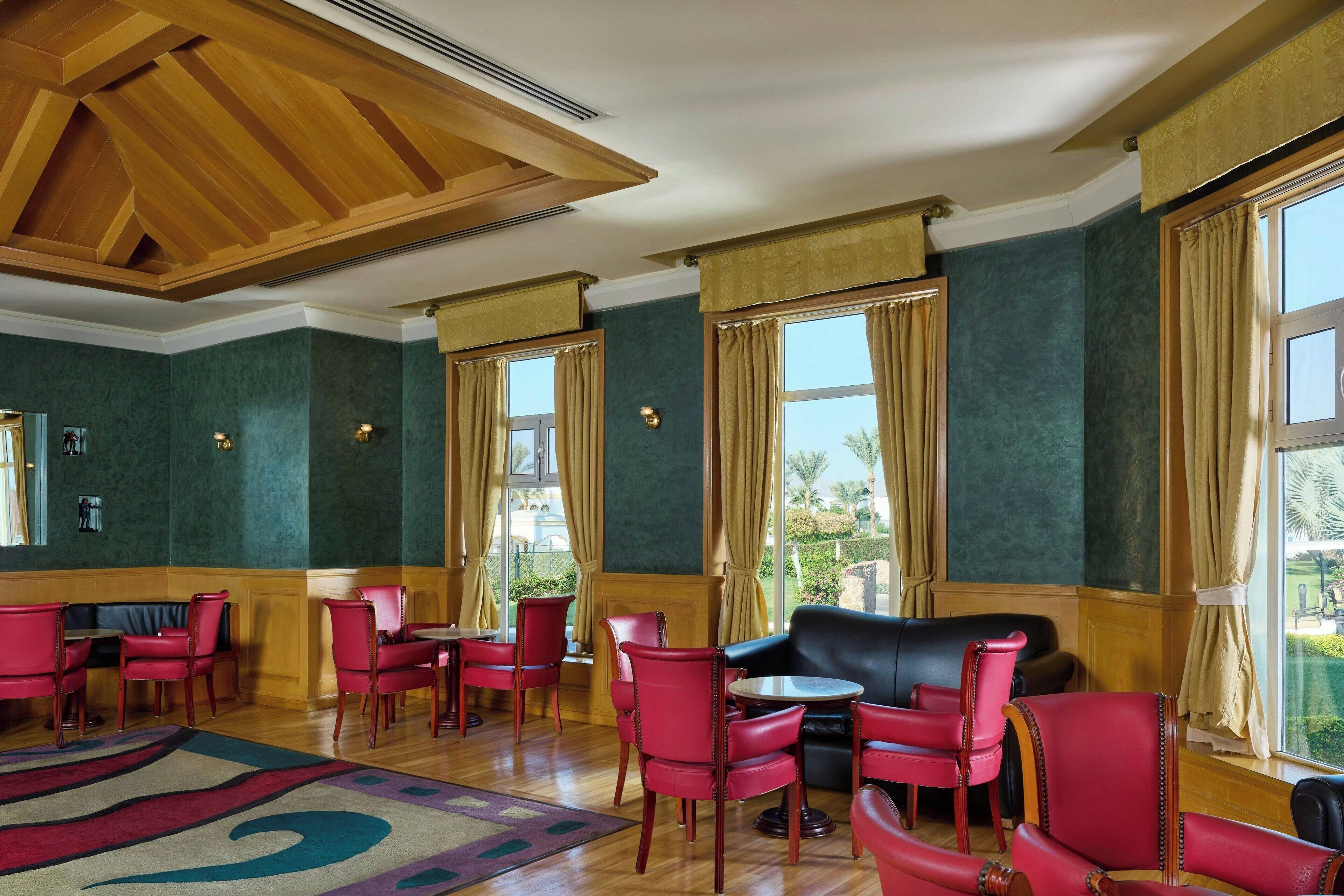 Un hall de hotel con paredes verdes, sillas rojas, un sofá negro y ventanas grandes con cortinas.