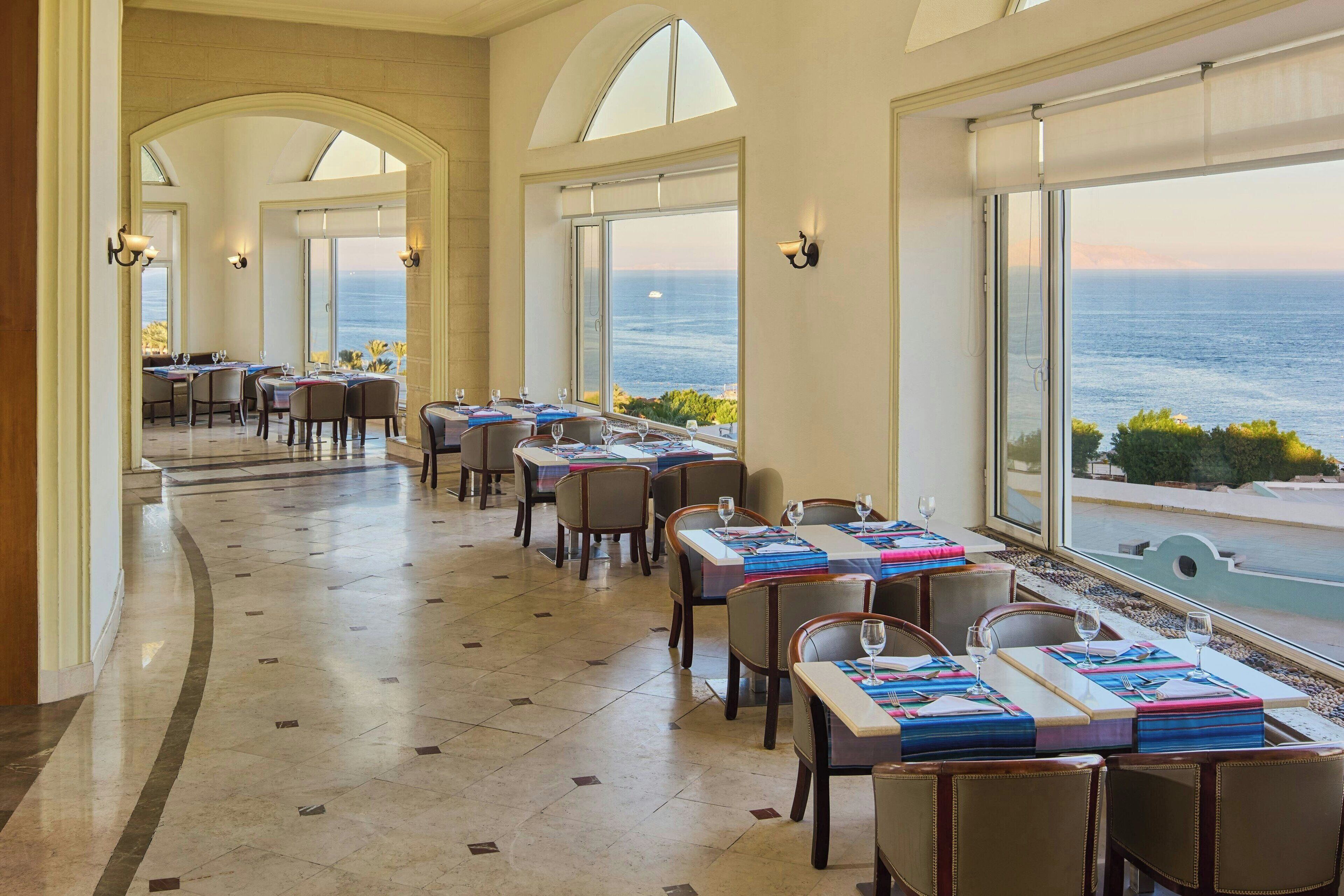 A restaurant with tables set for dining, large windows with ocean views, and a tiled floor.
