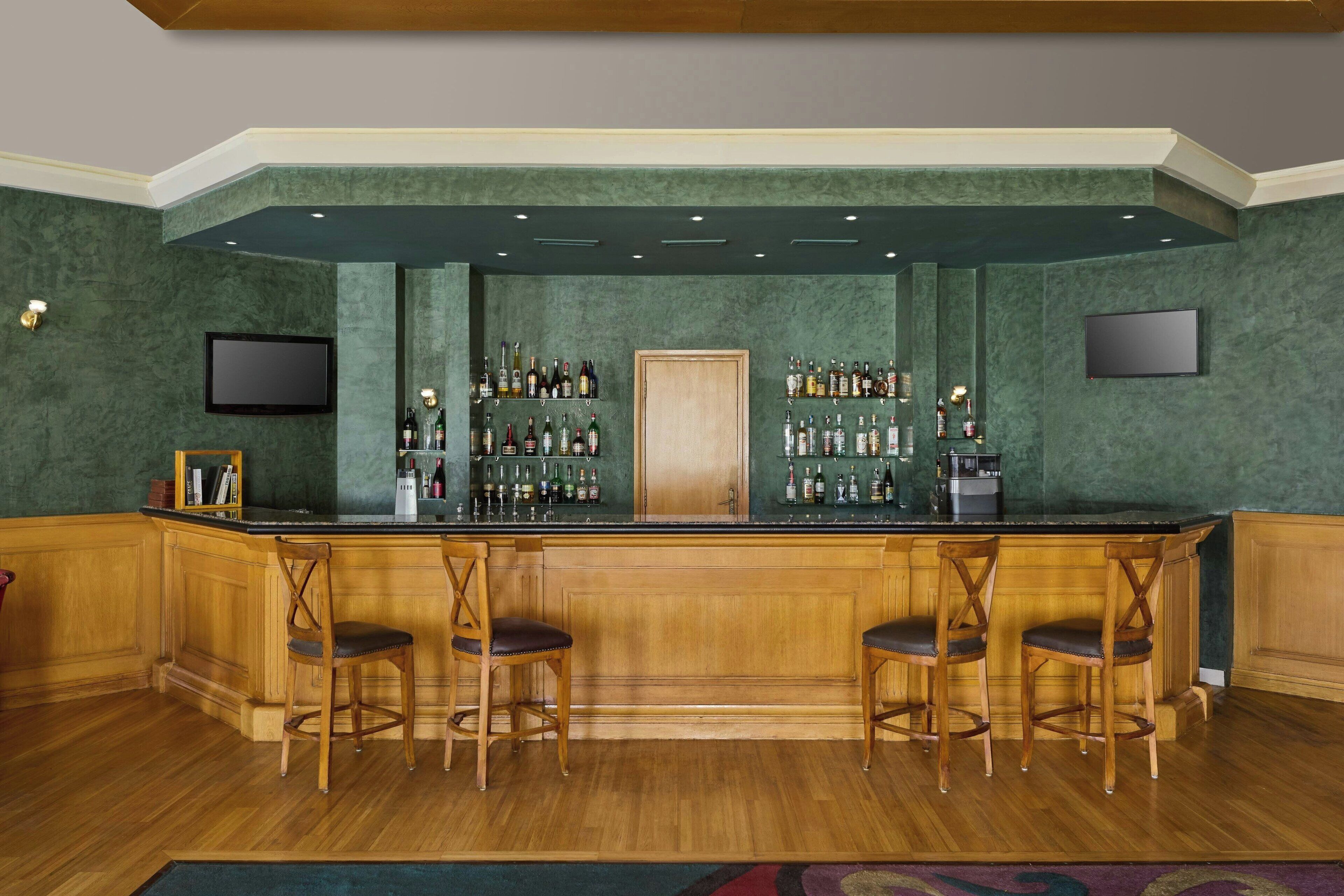 Una zona de bar con un mostrador de madera, dos taburetes y estantes llenos de botellas.