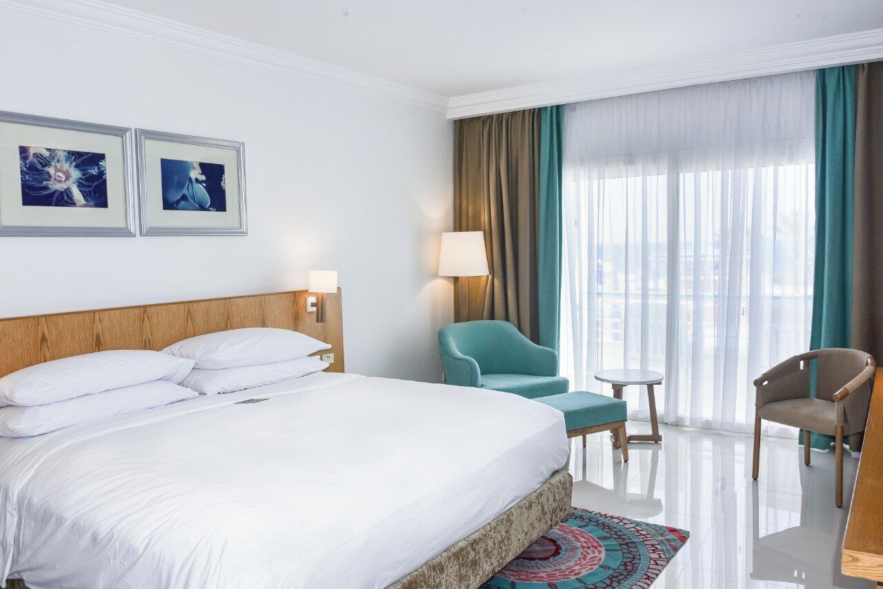 A hotel room with a large bed, two chairs, a small table, and a view of the city through the window.