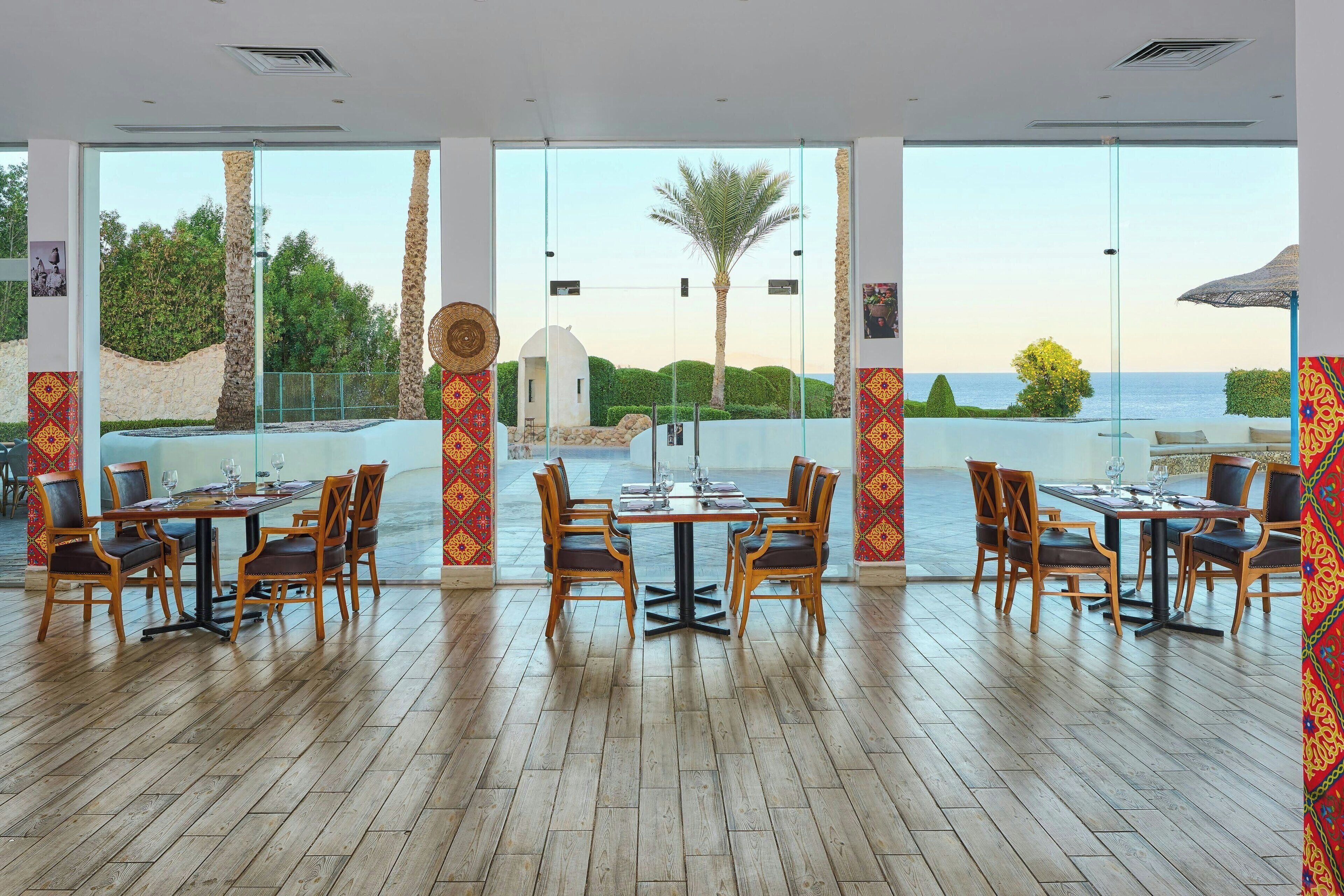 A dining area with wooden tables and chairs, large windows with a view of the sea and palm trees, and decorative wall panels.