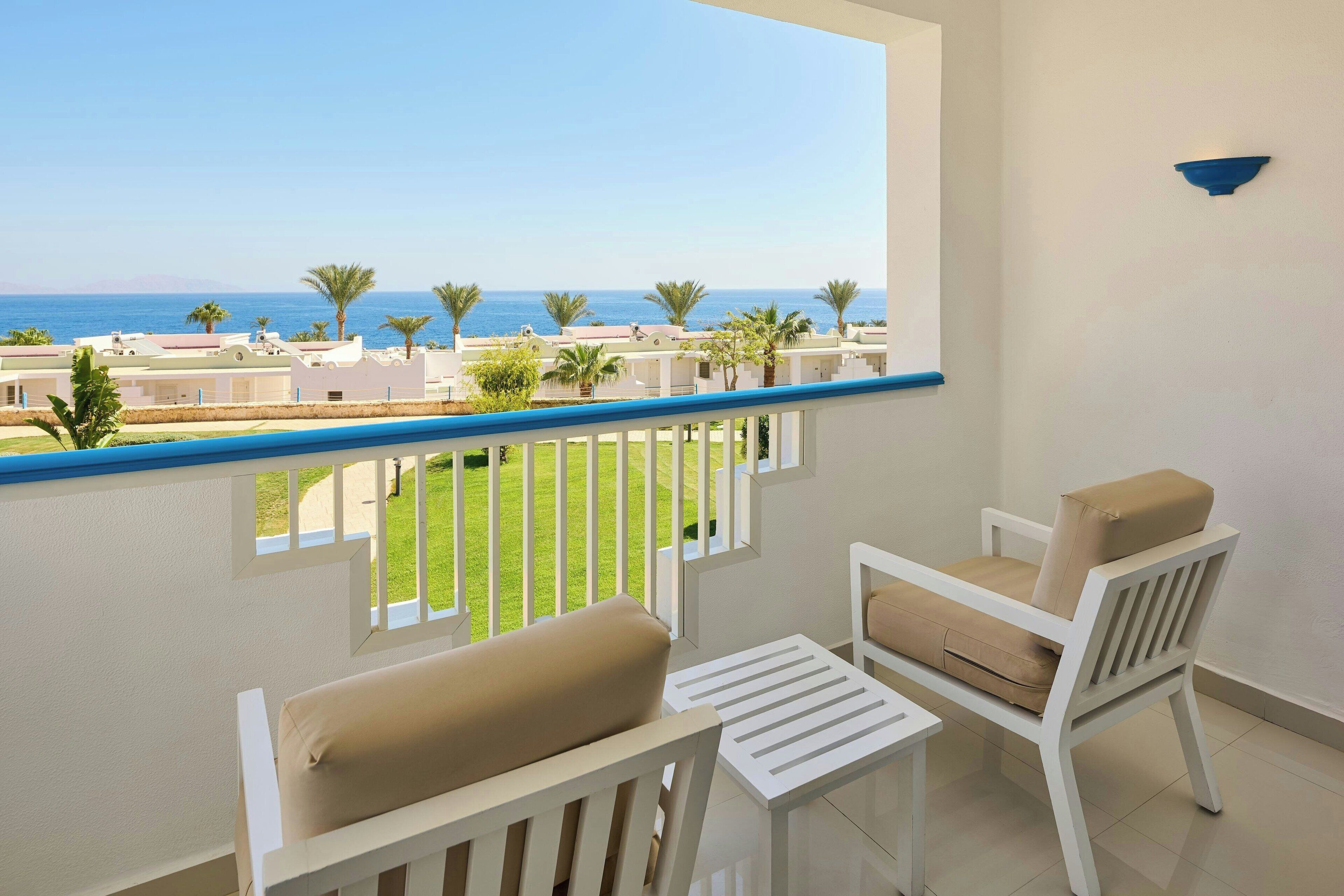 A balcony with a view of the sea, white furniture, and a blue railing.