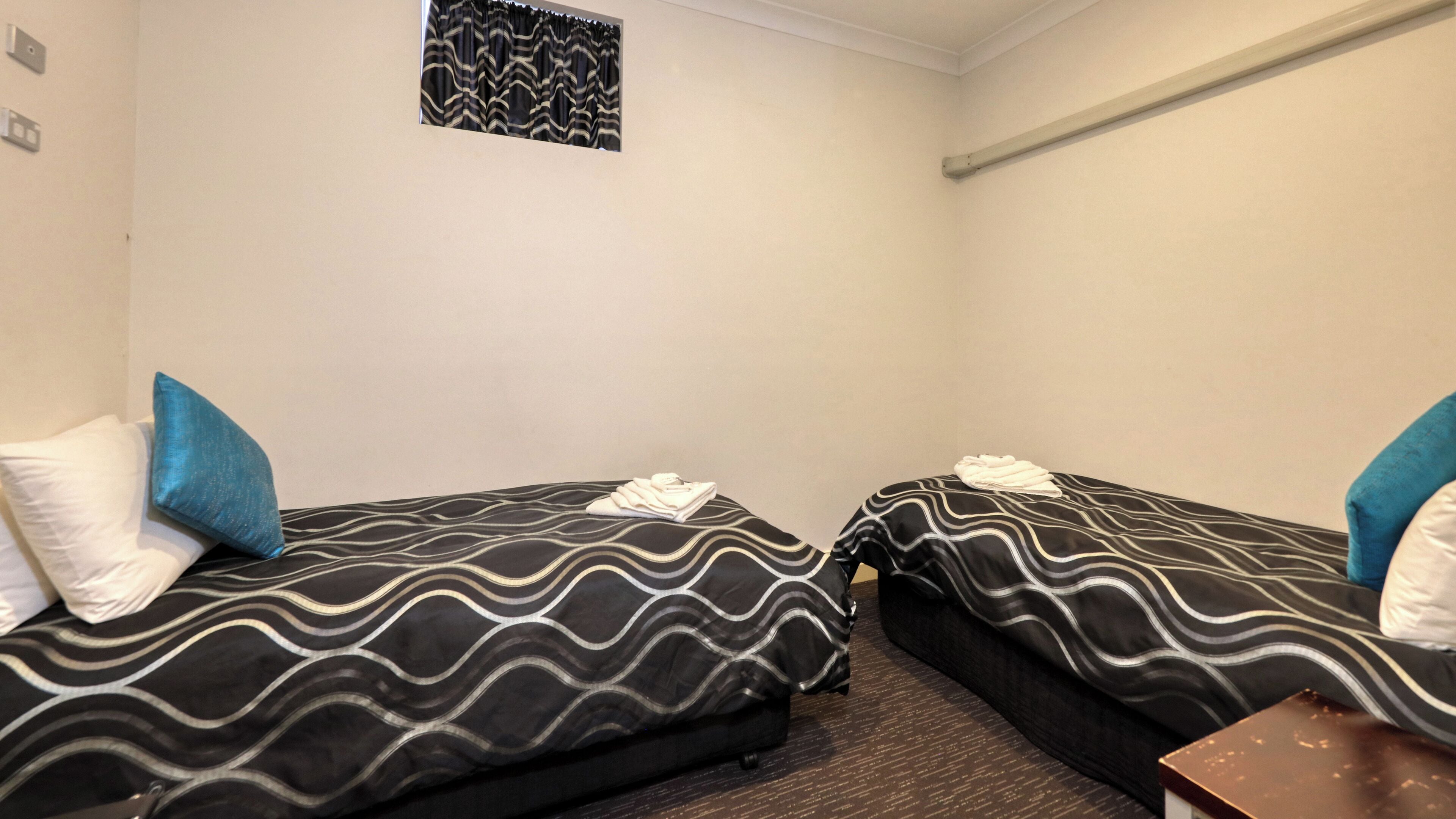 A small, compact room with two single beds, each with a patterned bedspread and pillows. There is a small table and a wall-mounted shelf.