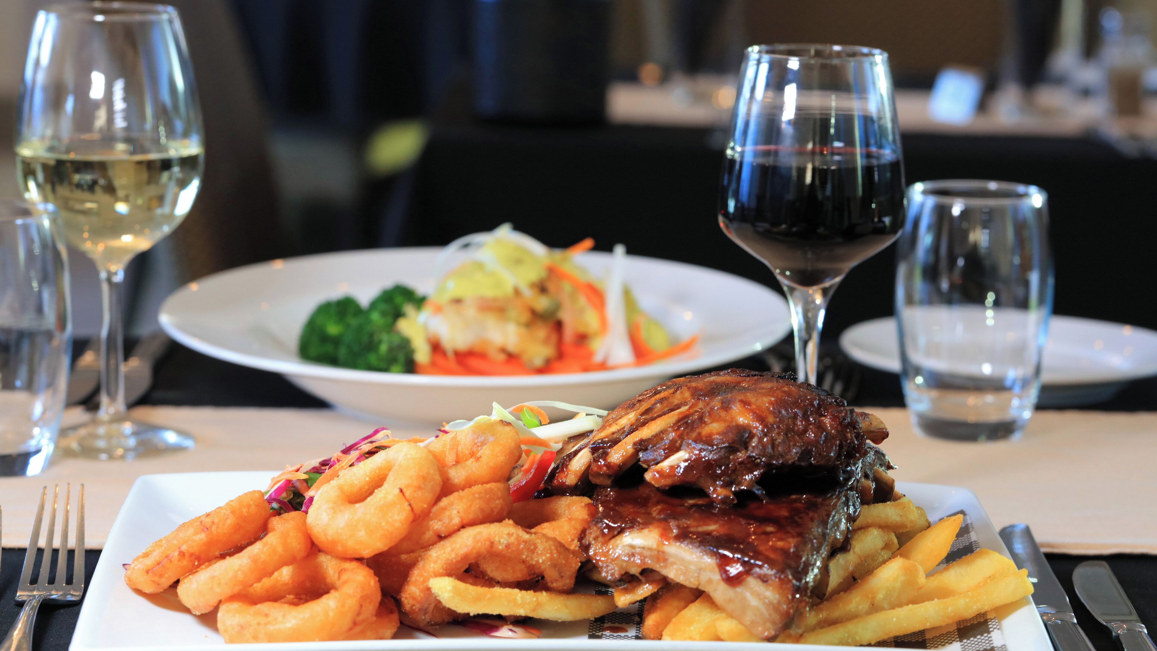 Un plato de costillas con aros de cebolla y papas fritas, una ensalada y dos copas de vino sobre una mesa.