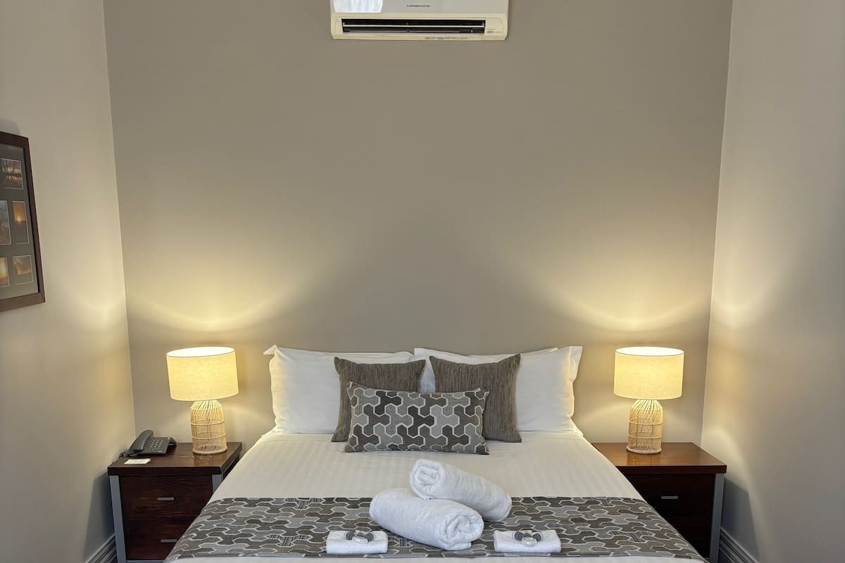 Ein ordentlich bezogenes Bett mit weißer Decke und grauen Kissen mit Muster, zwei Nachttische mit Lampen sowie eine Wand-Klimaanlage.