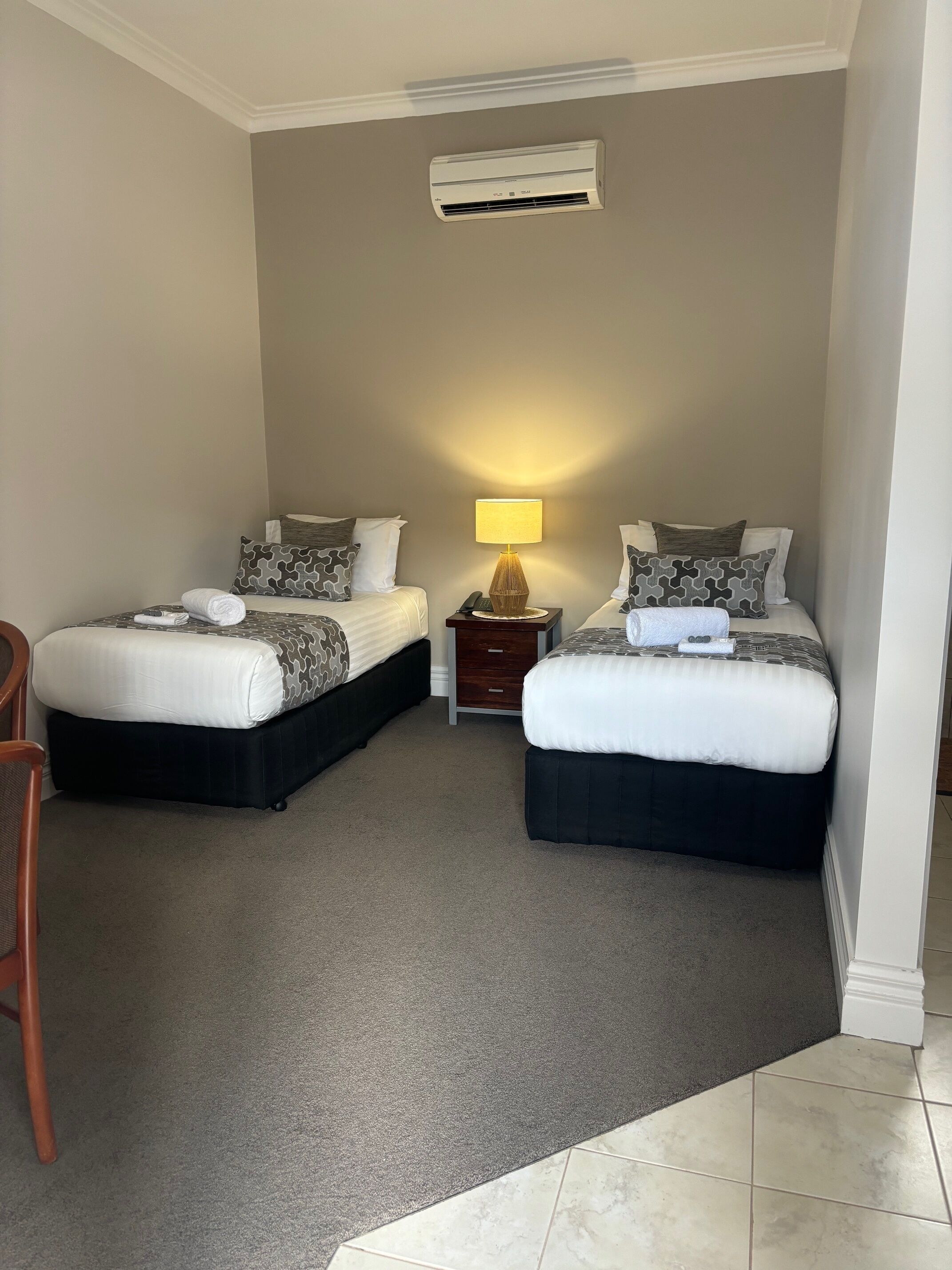 A hotel room with two beds, a nightstand, a lamp, and an air conditioning unit.