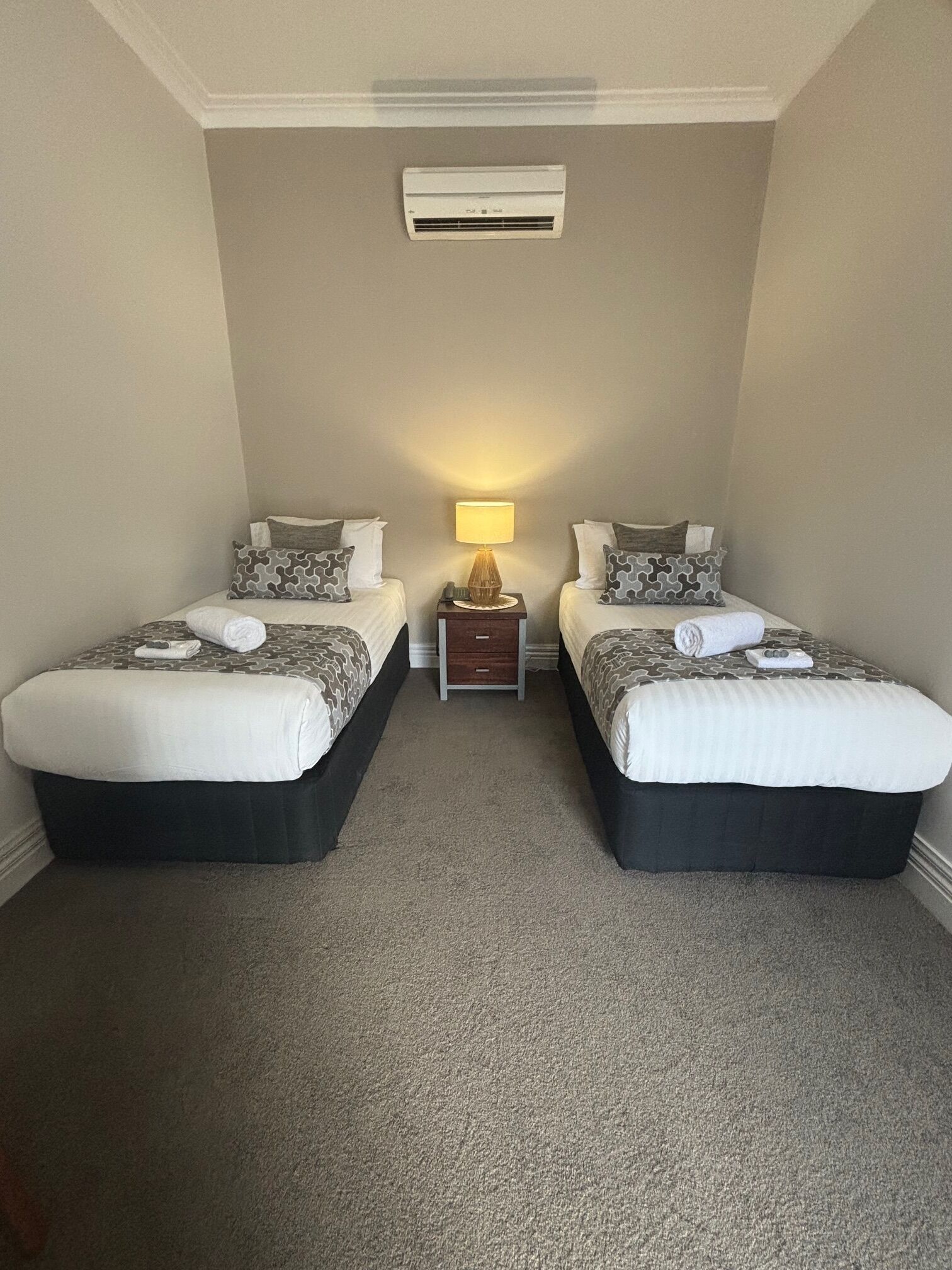 Quarto de hotel com duas camas, uma mesa de cabeceira com abajur e aparelho de ar condicionado na parede.