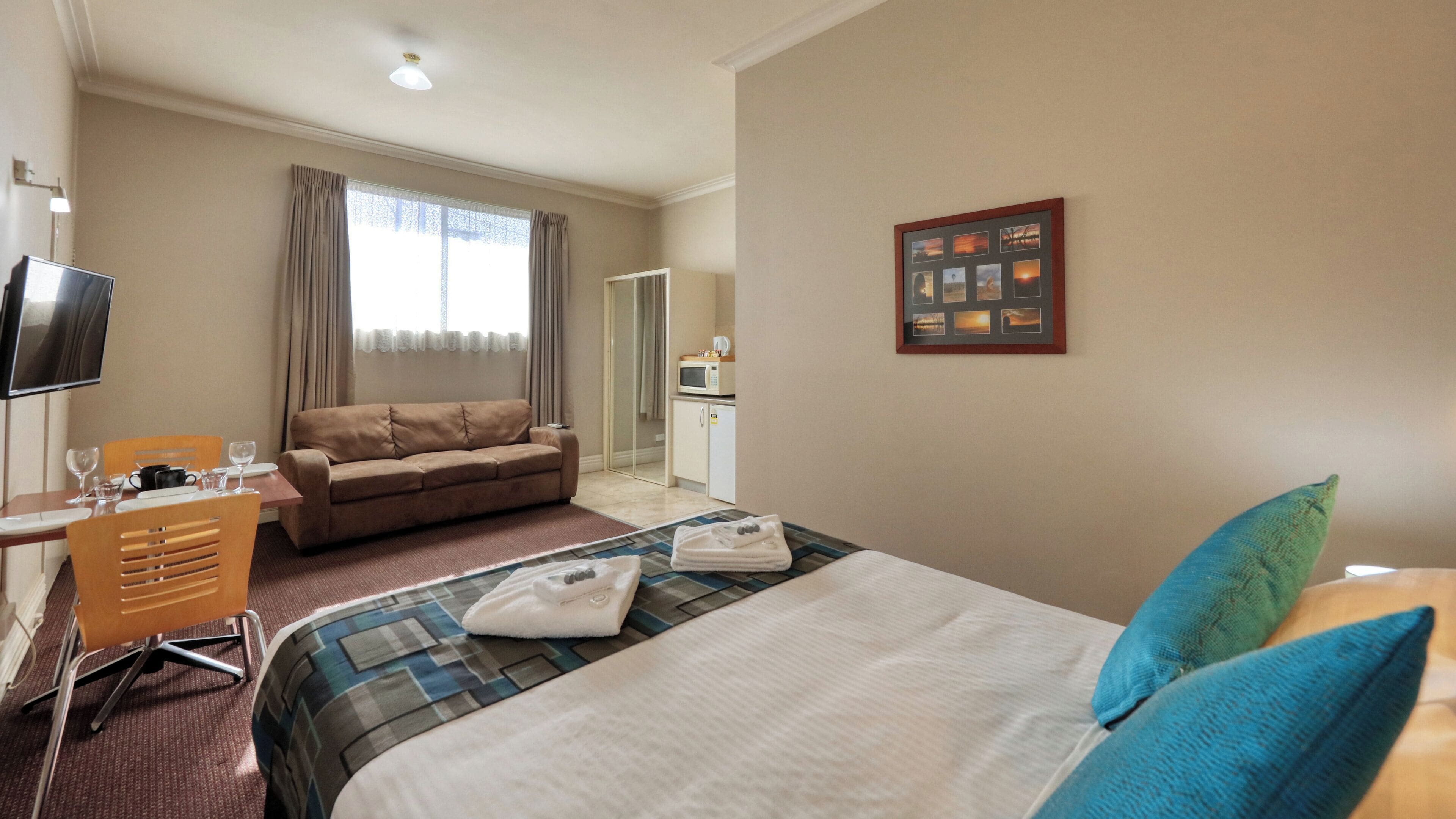 Habitación de hotel con cama, sofá, escritorio con silla, televisión, ventana con cortinas, microondas y un cuadro en la pared.