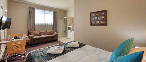 Habitación de hotel con cama, sofá, escritorio con silla, televisión, ventana con cortinas, microondas y un cuadro en la pared.