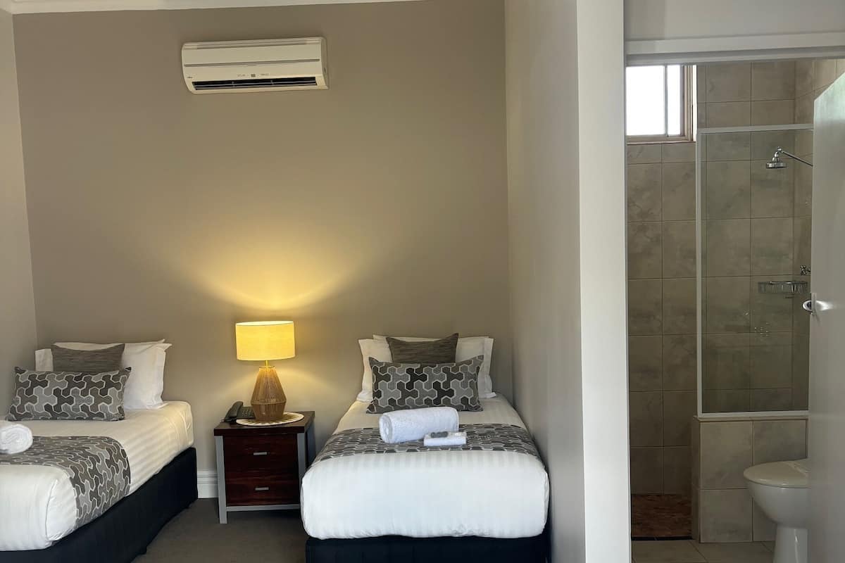 Ein Hotelzimmer mit zwei Betten, einem Nachttisch mit Lampe, einer Klimaanlage und einem Duschbereich.