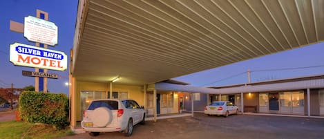 A motel with a covered parking area, a sign indicating no vacancy, and a car parked outside.