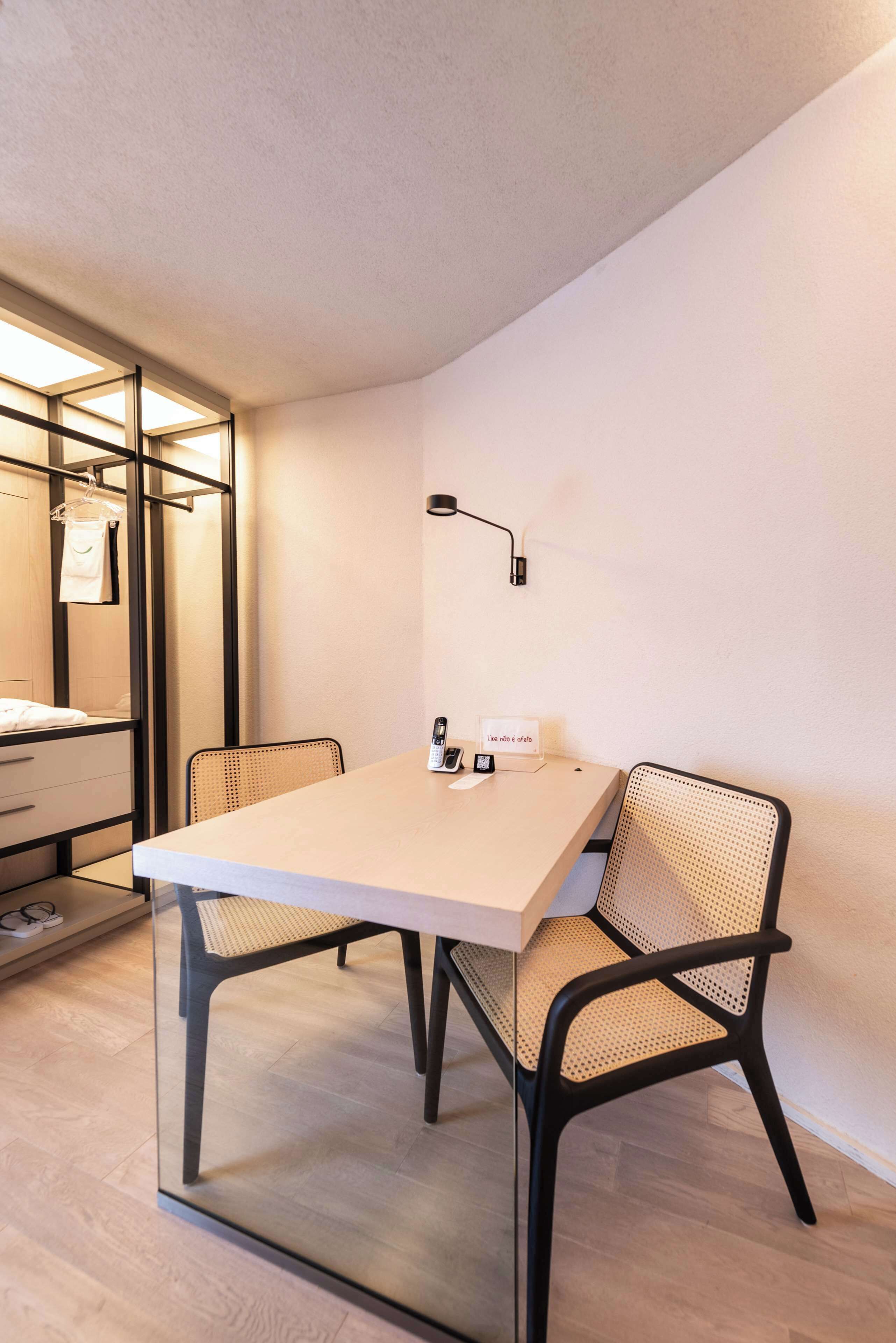 Una habitación moderna con separador de vidrio, una mesa de madera, dos sillas tejidas, una lámpara de pared y un letrero pequeño.