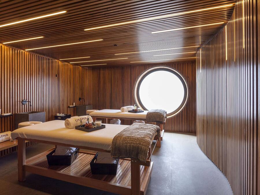 Une salle de spa moderne avec un lambris en bois, une table de massage, un lit de soins et une grande fenêtre circulaire.